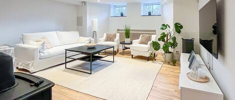 En moderne stue med sofa, sofabord og lænestole, et fjernsyn ophængt på væggen og en potteplante.