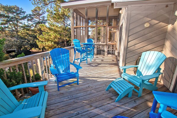 A wooden deck with blue Adirondack chairs and a table, overlooking a garden with trees and a house. - Soundside-Resort Amenities, Comm Pool-322, Out of Bounds (Corolla)