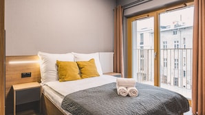 A hotel room with a bed, a nightstand, a window with curtains, and a view of buildings outside. - Calma Wanna Balkon Studio Centrum (Bydgoszcz)