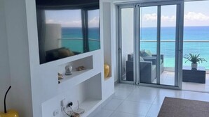 A modern living room with a large TV, white walls, and a glass door leading to a balcony with a view of the ocean. - A paradise in the Caribbean (Playa Juan Dolio)