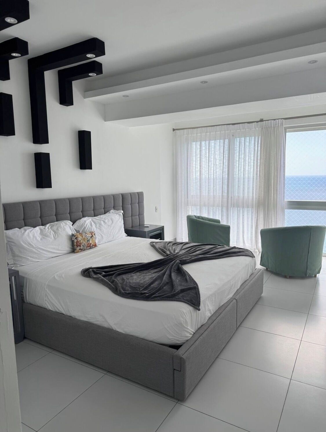 A modern bedroom with a large bed, a nightstand, a green armchair, and a view of the sea through sheer curtains.