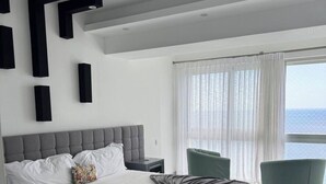 A modern bedroom with a large bed, a nightstand, a green armchair, and a view of the sea through sheer curtains. - A paradise in the Caribbean (Playa Juan Dolio)