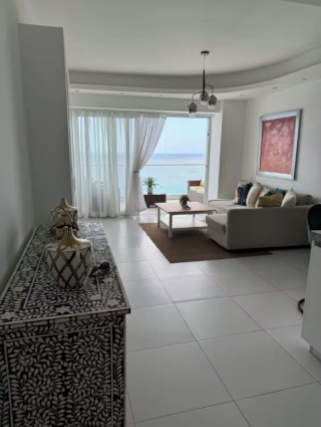 A modern living room with a sofa, coffee table, and decorative items. A large window offers a view of the sea.