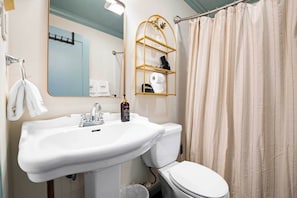 Un baño con un lavamanos blanco, un inodoro, una cortina de ducha y un estante con artículos de aseo.