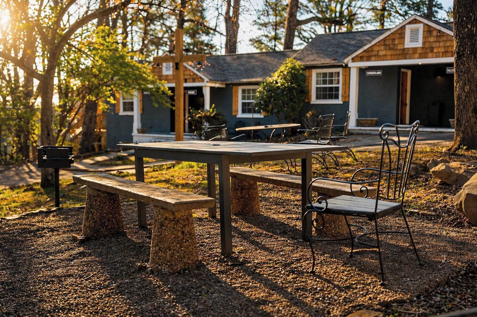 A rustic outdoor seating area with a table and benches, surrounded by trees and a building with a sign reading 'CINNAMON LAND'.