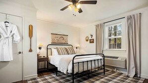Ein Schlafzimmer mit einem Bett, einem Nachttisch, einer Wandlampe, einem Deckenventilator, einem Fenster mit Vorhängen und einem Schachbrettmuster-Teppich.