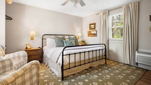 Een slaapkamer met een bed, een stoel, een nachtkastje, een plafondventilator, een raam met gordijnen en een radiator.