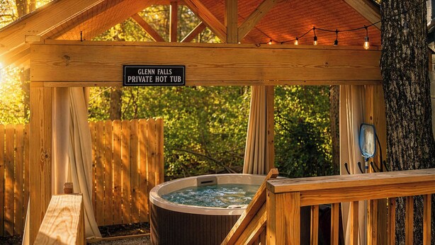 Een houten jacuzzi met een bordje waarop staat 'Glenn Falls Private Hot Tub', omringd door een houten terras en een overdekt gebied met lichtslingers.