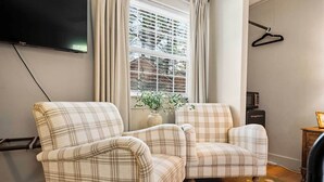 Een woonkamer met twee geruite fauteuils, een flatscreen televisie, een raam met zonwering, een hangende kapstok en een magnetron op een houten kast.