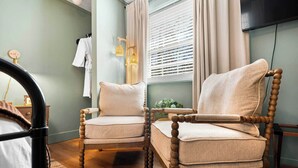 Una habitación con dos sillas de madera, una cama, un ventanal con persianas, una mesita con una planta y un televisor instalado en la pared.
