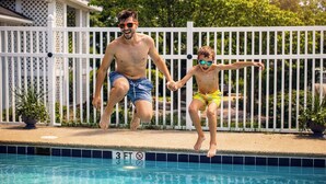 Dos personas saltando a una piscina con una cerca blanca y una casa al fondo.