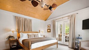 Une chambre avec un lit en bois, un ventilateur de plafond, une table de chevet avec une lampe et une vue sur l’extérieur, visible à travers une porte vitrée.