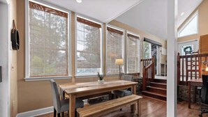 Un comedor con una mesa de madera y bancos, ventanas grandes con persianas, una lámpara y una escalera que sube a otro nivel.