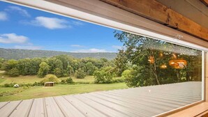 Une vue depuis une fenêtre, avec un toit en métal, donnant sur un paysage verdoyant parsemé d’arbres et sur une petite construction.