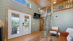 Un intérieur chaleureux avec une chambre à coucher située en mezzanine, un coin repas avec une table et des chaises, et un salon avec un canapé et un téléviseur affichant le nom « QUAIL RUN FARM ».