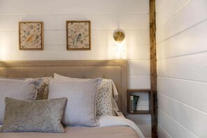 A bedroom with a bed, pillows, a headboard, two framed floral artworks, a wall-mounted light fixture, and a small wooden side table with a book.