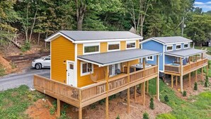 A two-story wooden cabin with a deck, a car parked nearby, and a forested backdrop. - The Honeycomb Cabin (Chattanooga)