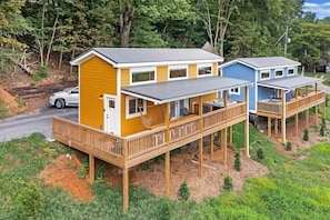 A two-story wooden cabin with a deck, a car parked nearby, and a forested backdrop.