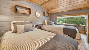 Ein schlicht eingerichtetes Schlafzimmer im Blockhausstil mit zwei Betten, einer Holzbalkendecke, einem Fenster mit Blick und einem Bücherregal.