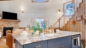 A modern kitchen with a stone fireplace, a large island with a sink, and a flat-screen TV mounted above the fireplace. - Lookout Silo with Mountain Views & Hot Tub (Chattanooga)