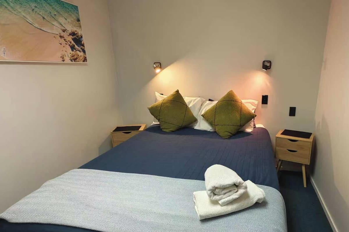 Una cama bien hecha con una colcha azul, dos almohadas verdes y una toalla blanca doblada en la cabecera. Hay dos mesitas de noche con lámparas y una luz empotrada en la pared.