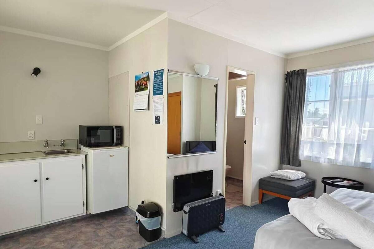 Una habitación de hotel con kitchenette, una cama, una mesita con una silla, una ventana con cortinas y una puerta que da a otra habitación.