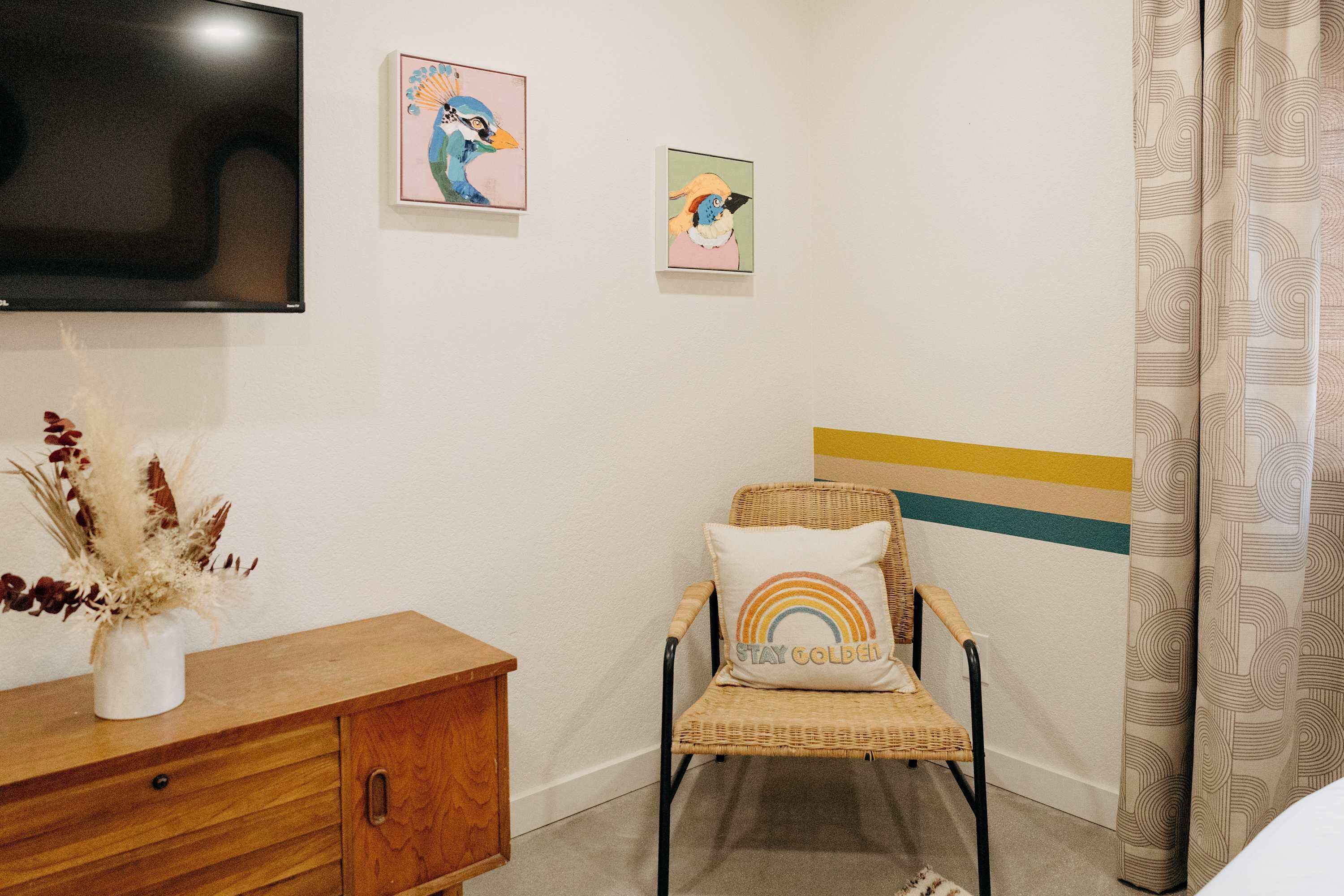 A room with a television mounted on the wall, a wicker chair with a cushion, a wooden cabinet, and a wall with two framed artworks.