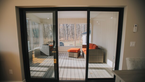 A room with a sliding glass door leading to an outdoor deck with a checkered floor, wicker furniture, and a view of trees. - The Creek House on Kaaterskill (Palenville)