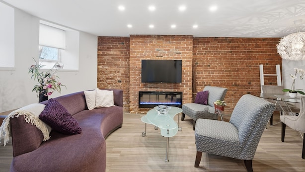 A living room with a brick wall, a flat-screen TV, a fireplace, a sofa, a chair, and a glass coffee table. - Modern Heritage Apartment in the Heart of Halifax (Halifax)