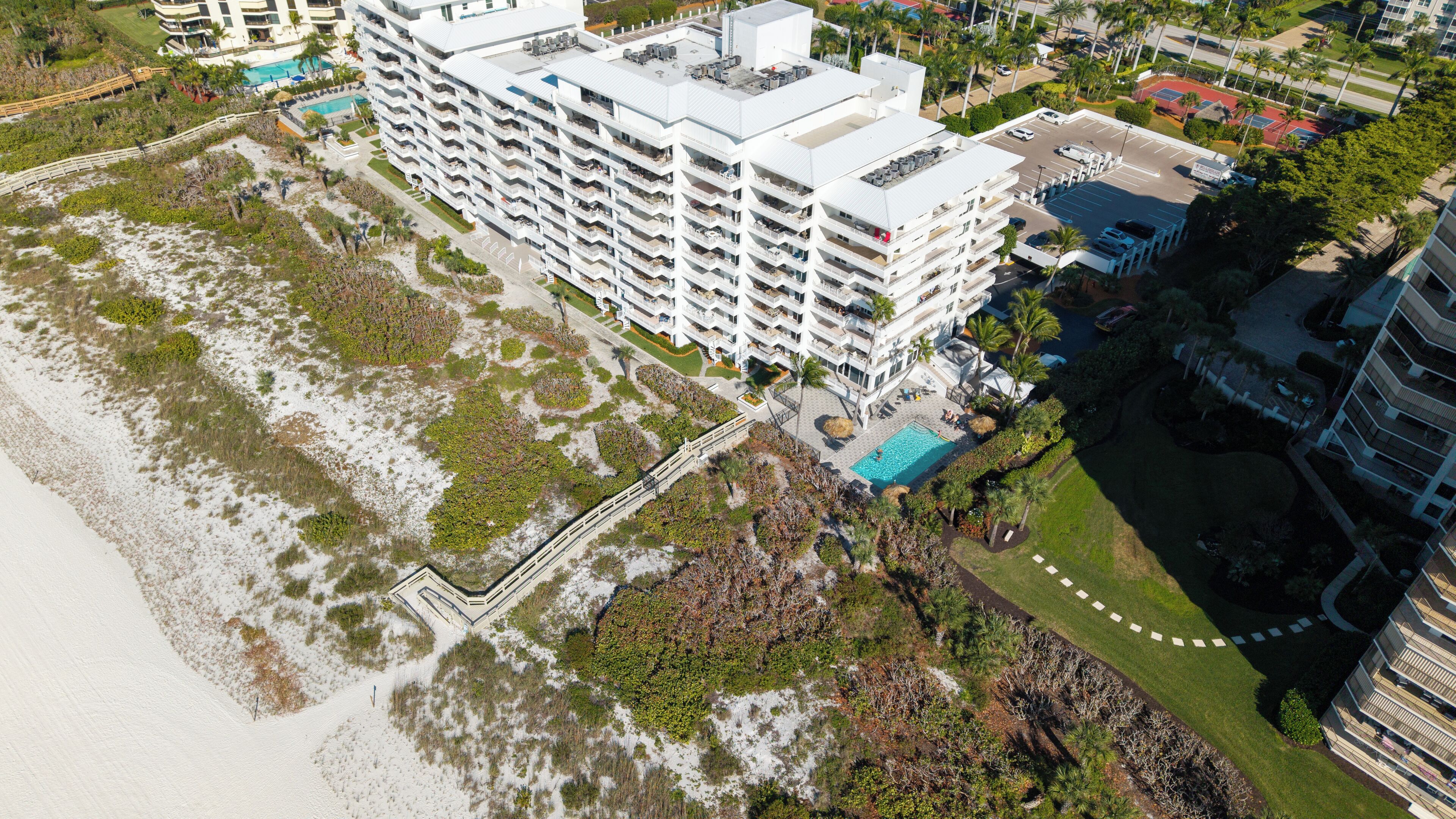 A multi-story hotel with a rooftop pool, adjacent to a beach with sandy areas and sparse vegetation.