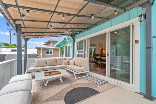 A covered patio area with a sectional sofa, a coffee table, and a dining area visible through glass sliding doors.