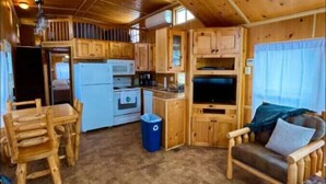 A cozy cabin interior with wooden walls and floors, a dining area with a table and chairs, a kitchen with appliances, and a seating area with a couch and pillows. - Little Lantern Cabin (Indianapolis)