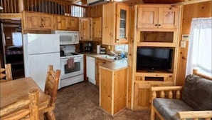 A cabin interior with wooden walls and ceiling, a kitchen area with a stove, refrigerator, and cabinets, a dining table with chairs, and a sofa.