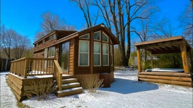A wooden cabin with a deck and a covered area, surrounded by snow and trees.