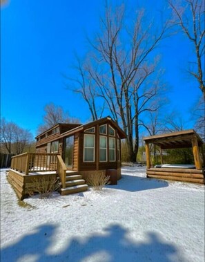 Una cabaña de madera con un balcón y un espacio cubierto, rodeada de nieve y árboles.