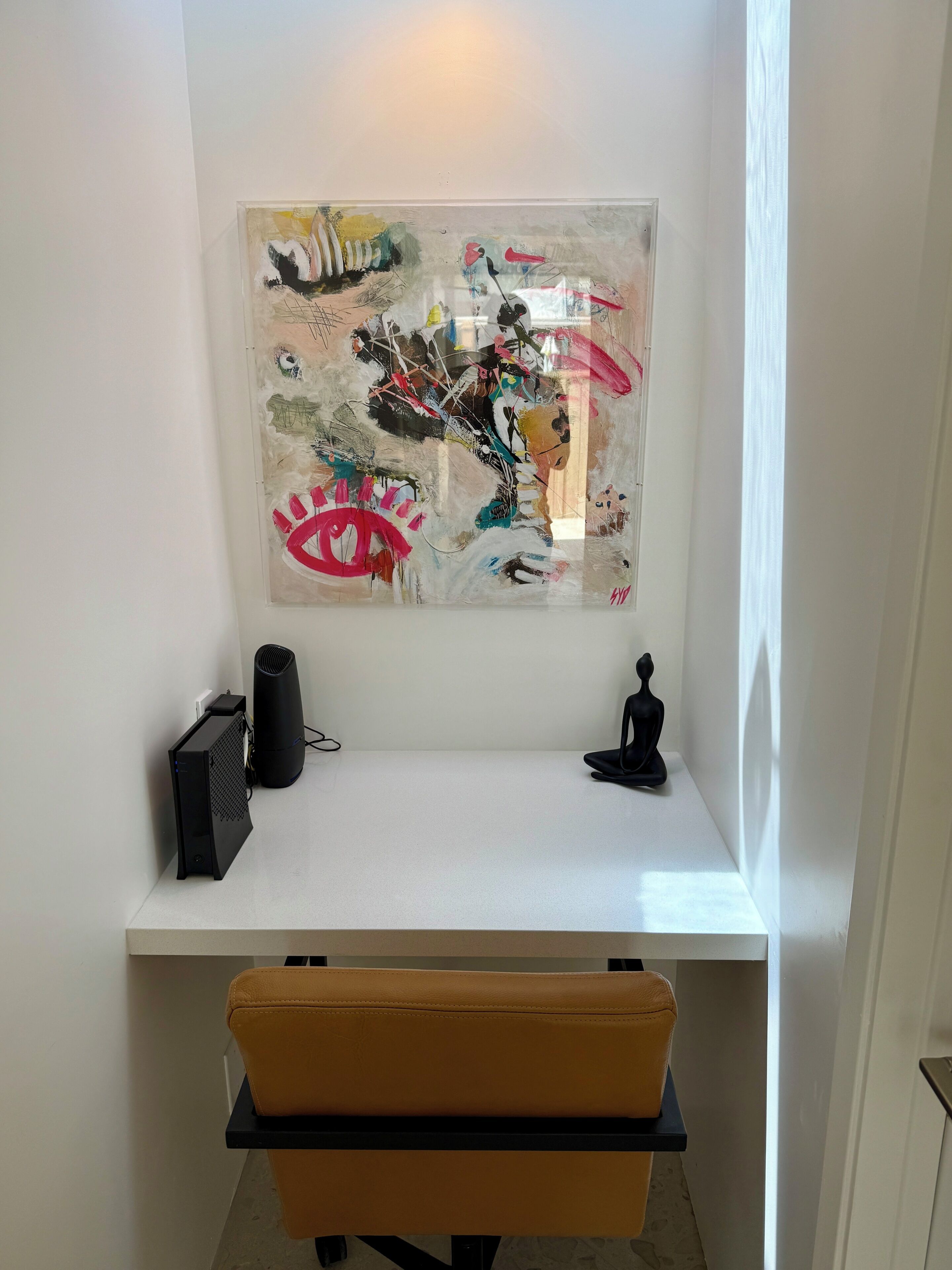 Ein weißer Schreibtisch mit einem Computer, einem schwarzen Lautsprecher und einer schwarzen Skulptur. Darüber an der Wand hängt ein farbenfrohes abstraktes Gemälde.