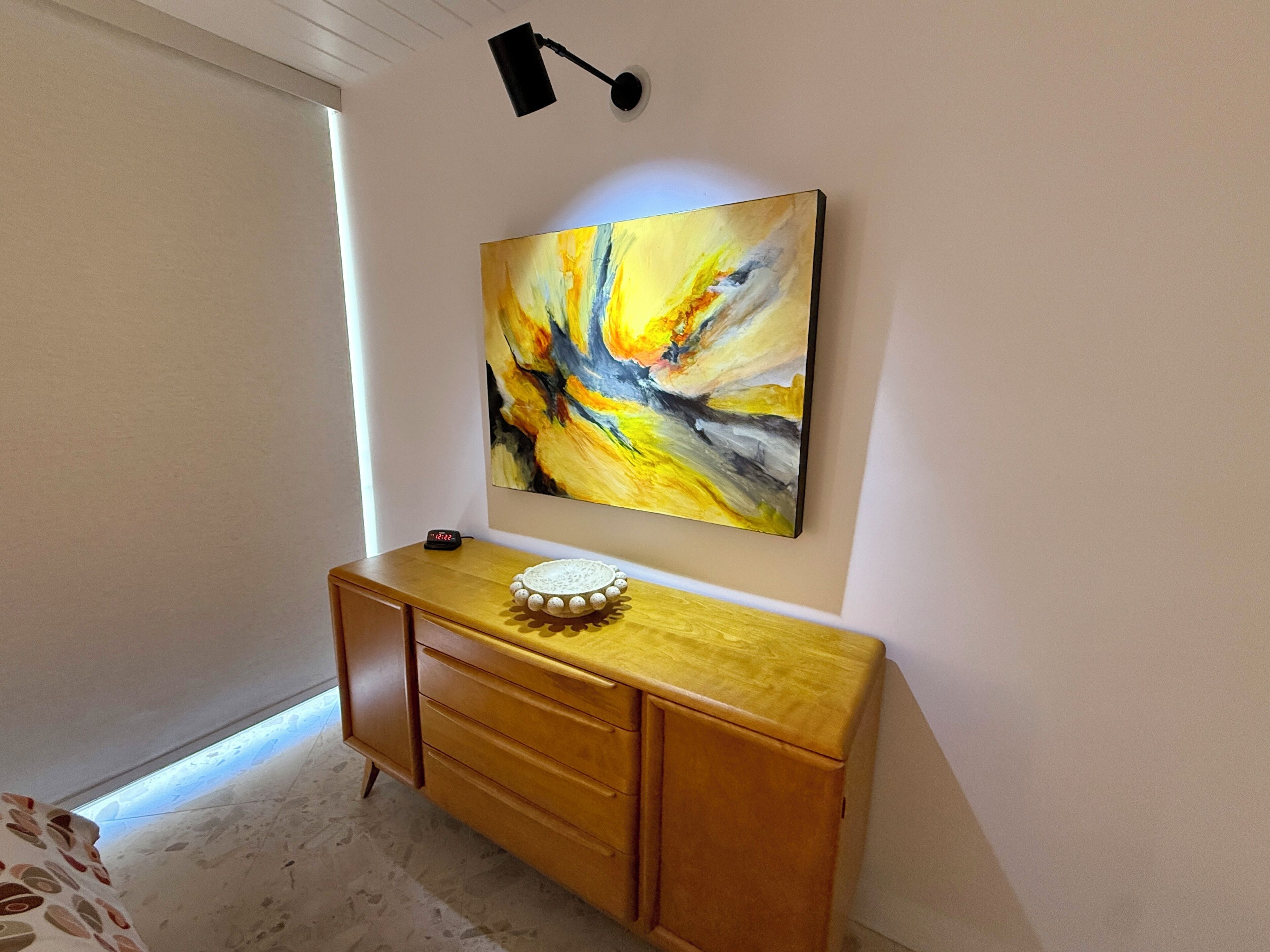 Quarto com escrivaninha de madeira, quadro abstrato pendurado na parede e uma pequena mesa com um objeto decorativo.