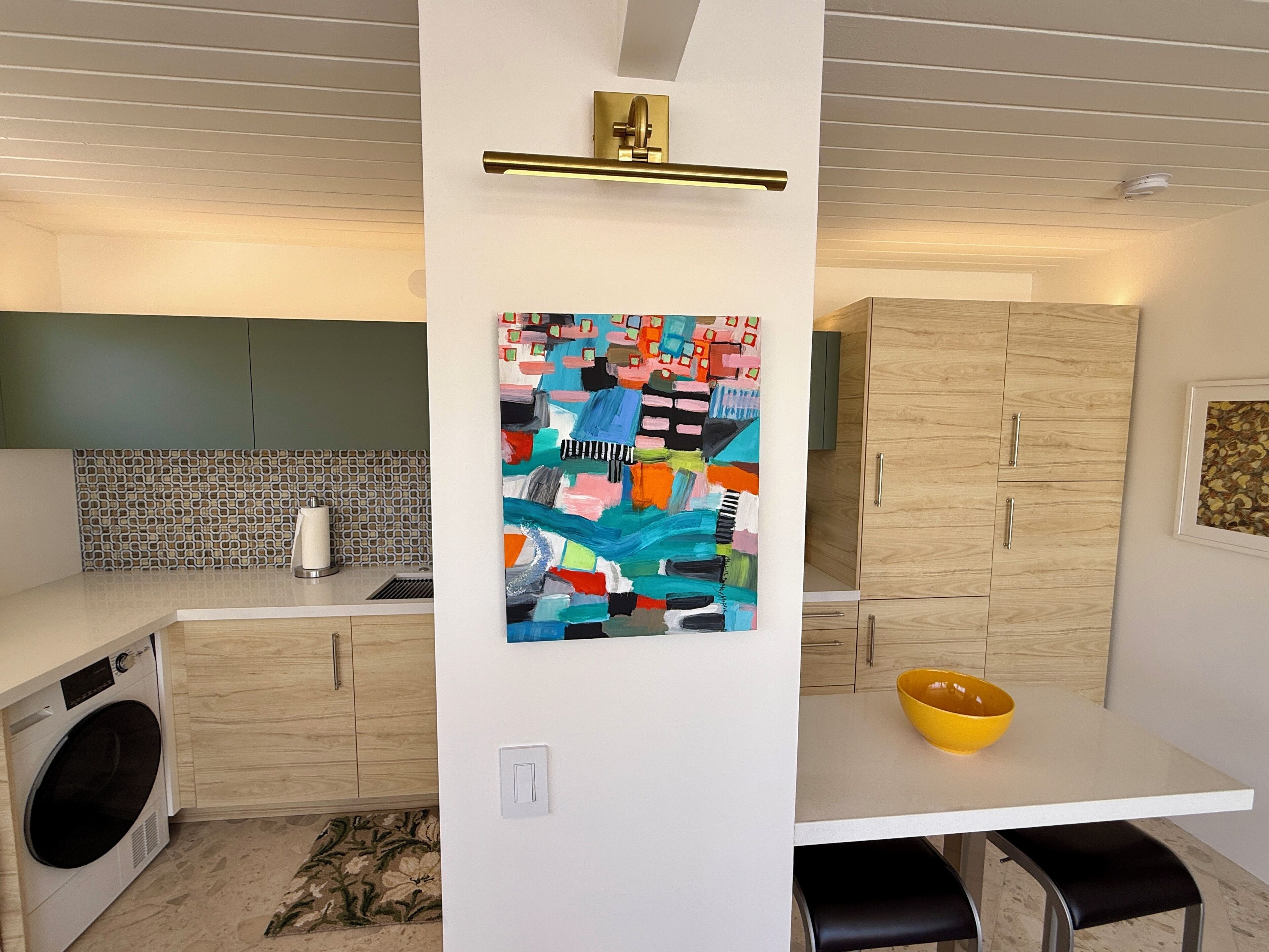 A modern kitchen with a central island, a washing machine, and a colorful abstract painting.