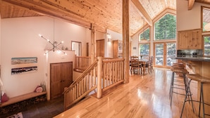Un intérieur spacieux en bois, avec un plafond haut, un escalier, un coin repas et de grandes fenêtres offrant une vue sur l’extérieur.