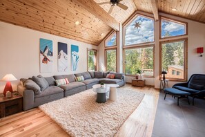 A modern living room with a sectional sofa, a round coffee table, and large windows offering a view of the outdoors. - Vibrance at Alpine by Tahoe Getaways - Ski Shuttle - 4BR w Hot Tub & Sauna (Alpine Meadows)