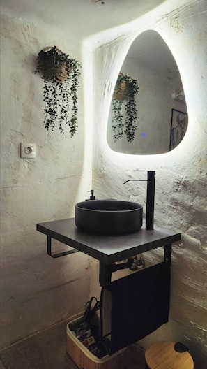 Un baño con un lavabo redondo, un espejo y plantas colgantes.