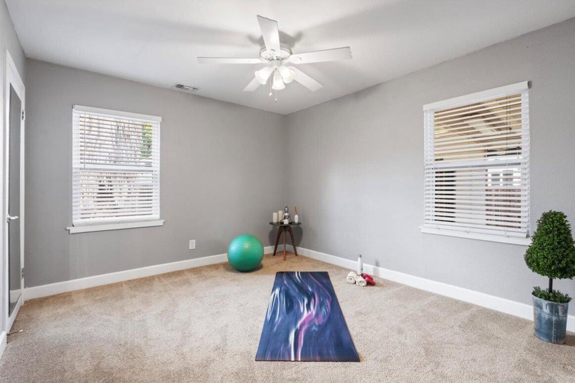Una habitación con ventilador de techo, dos ventanas con persianas, una esterilla de yoga, una pelota de ejercicios verde y una pequeña planta en maceta.