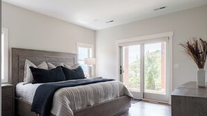 A modern bedroom with a large bed, a nightstand, a bedside table, a lamp, and a view through a sliding glass door. - Luxe | 8BR 6 Kings | Sleeps 22 | 2HotTub | Firepit (Banner Elk)
