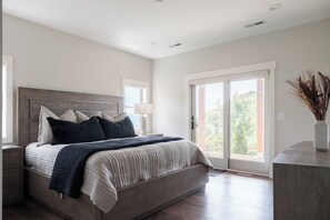 A modern bedroom with a large bed, a nightstand, a bedside table, a lamp, and a view through a sliding glass door. - Luxe | 8BR 6 Kings | Sleeps 22 | 2HotTub | Firepit (Banner Elk)