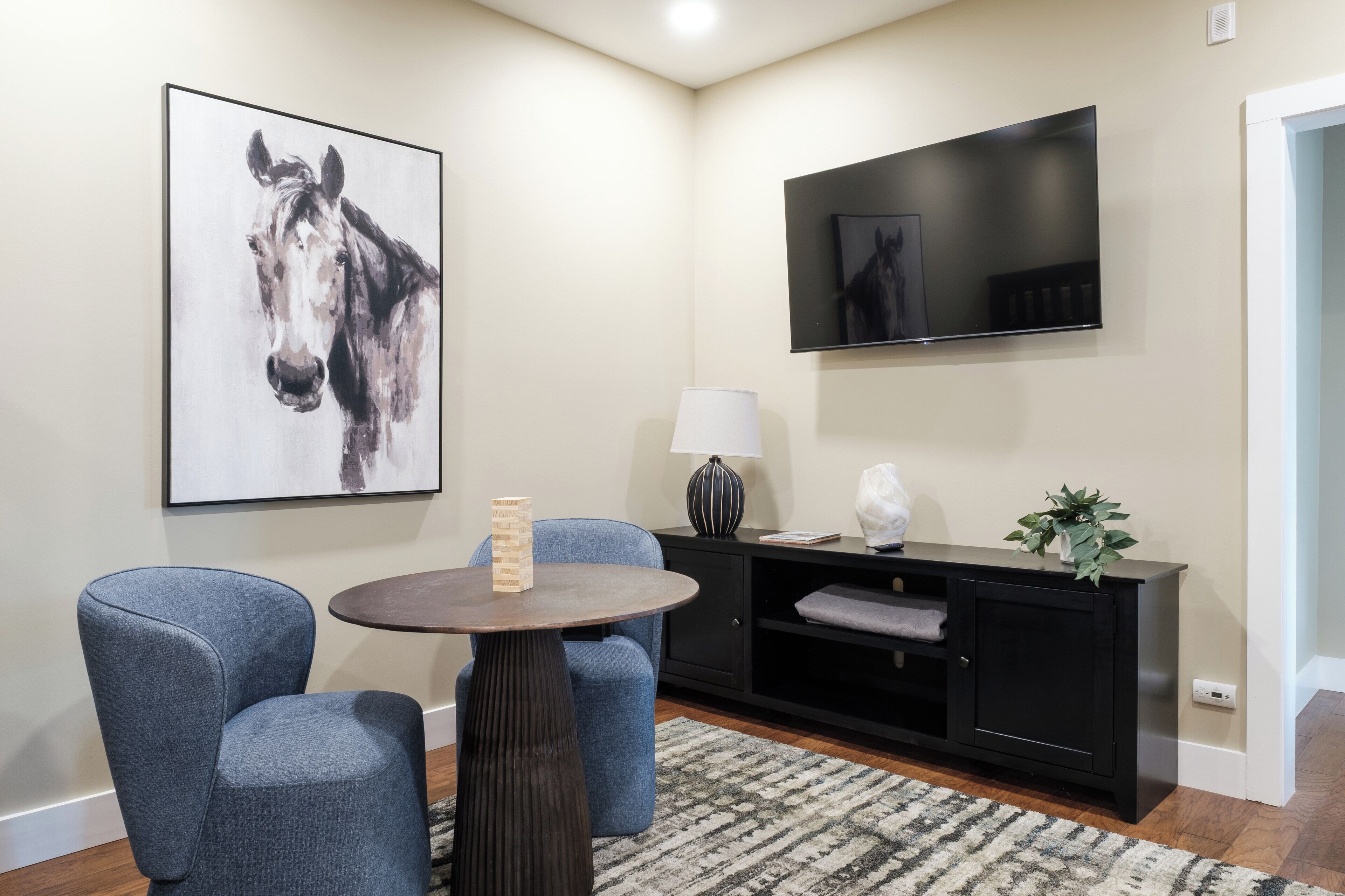A modern living room with a round table, two blue chairs, a black entertainment unit, a mounted TV, and a large horse painting on the wall.