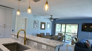 Una cocina con encimera de mármol, un lavabo y un ventilador de techo. Hay una zona de comedor con mesa y sillas, y una sala de estar con sofá y televisor.