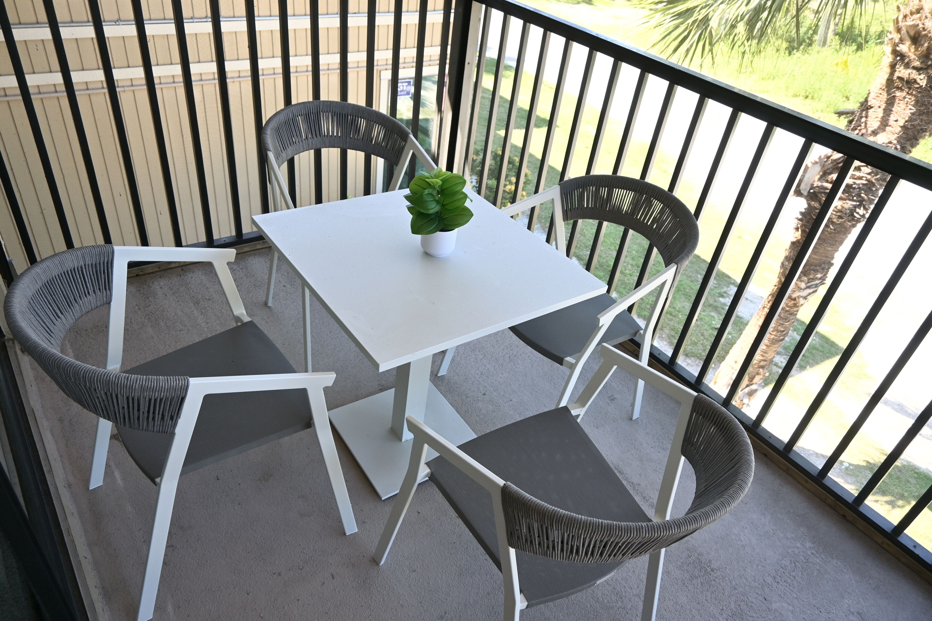 A balcony with a white table and four wicker chairs, a small potted plant, and a view of a palm tree and grass.