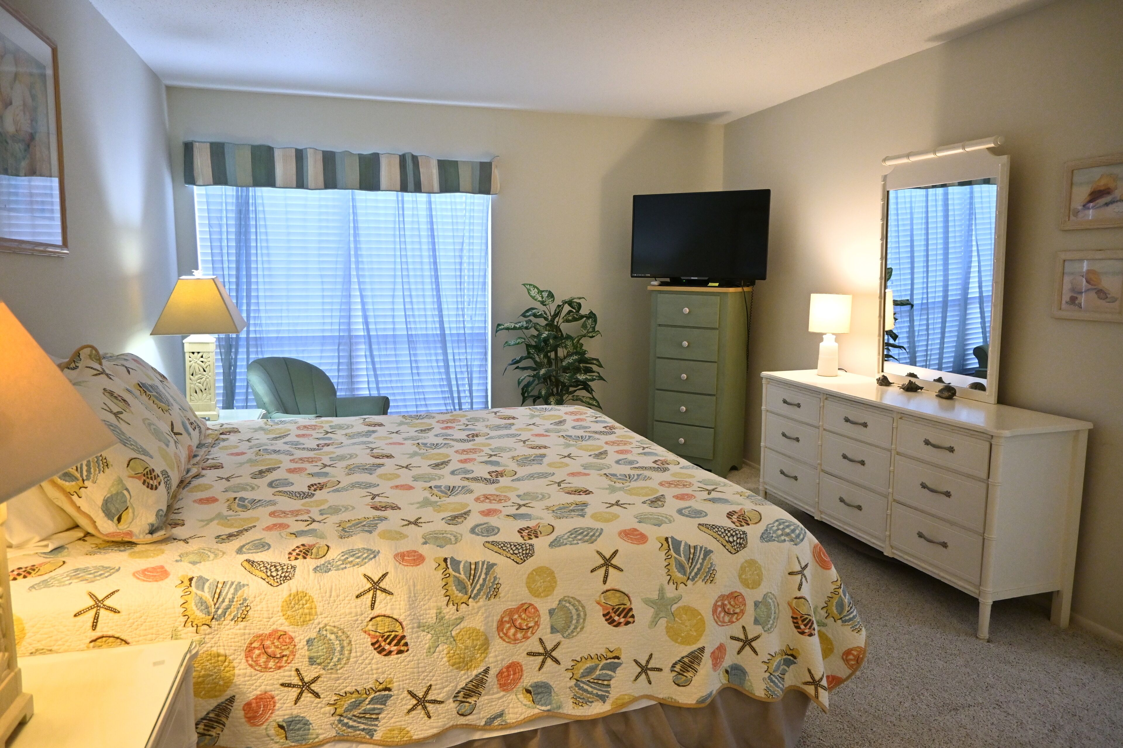 A bedroom with a bed, a dresser, a television, a lamp, and a window with curtains.