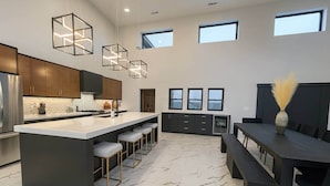 A modern kitchen with a central island, bar stools, and pendant lighting. There are three windows providing natural light. - Pure MUCHigan (Holland)