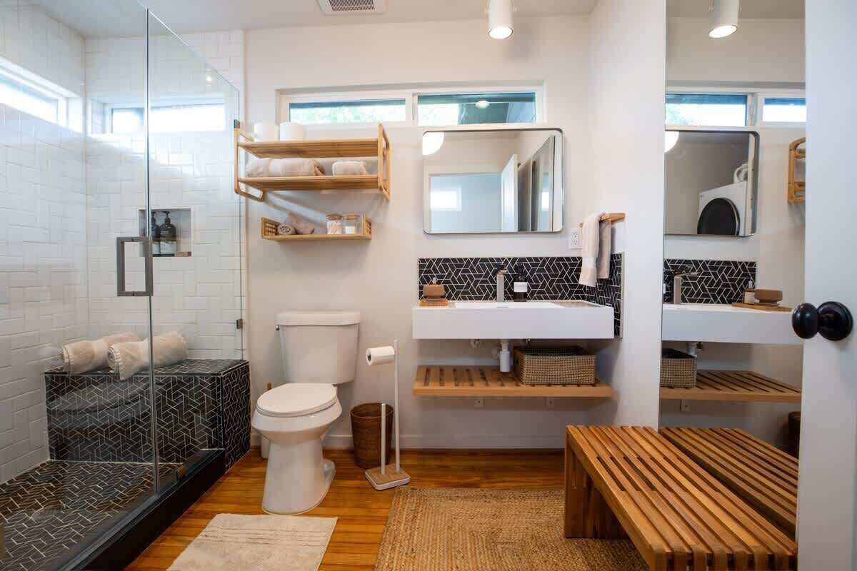 A modern bathroom with a glass shower enclosure, a white toilet, a wooden bench, and a sink with a mirror above it.